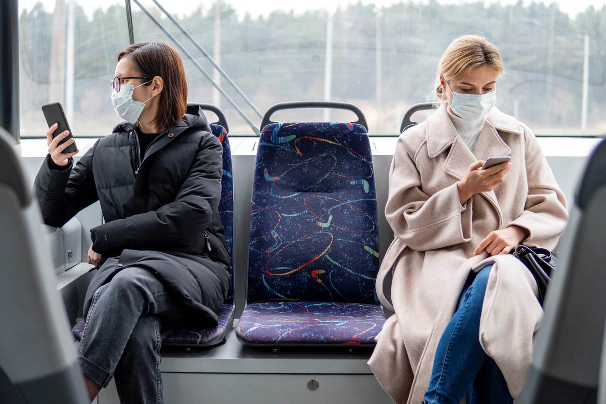 Twee vrouwen in een bus met een mondkapje op op hun telefoon en 1,5 meter afstand tussen hen