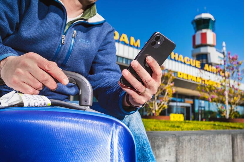 Zittende man met koffer en telefoon in zijn hand voor Rotterdam Airport