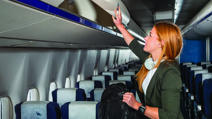 Vrouw in vliegtuig met smartwatch en koptelefoon om die iets uit het bagagevak pakt